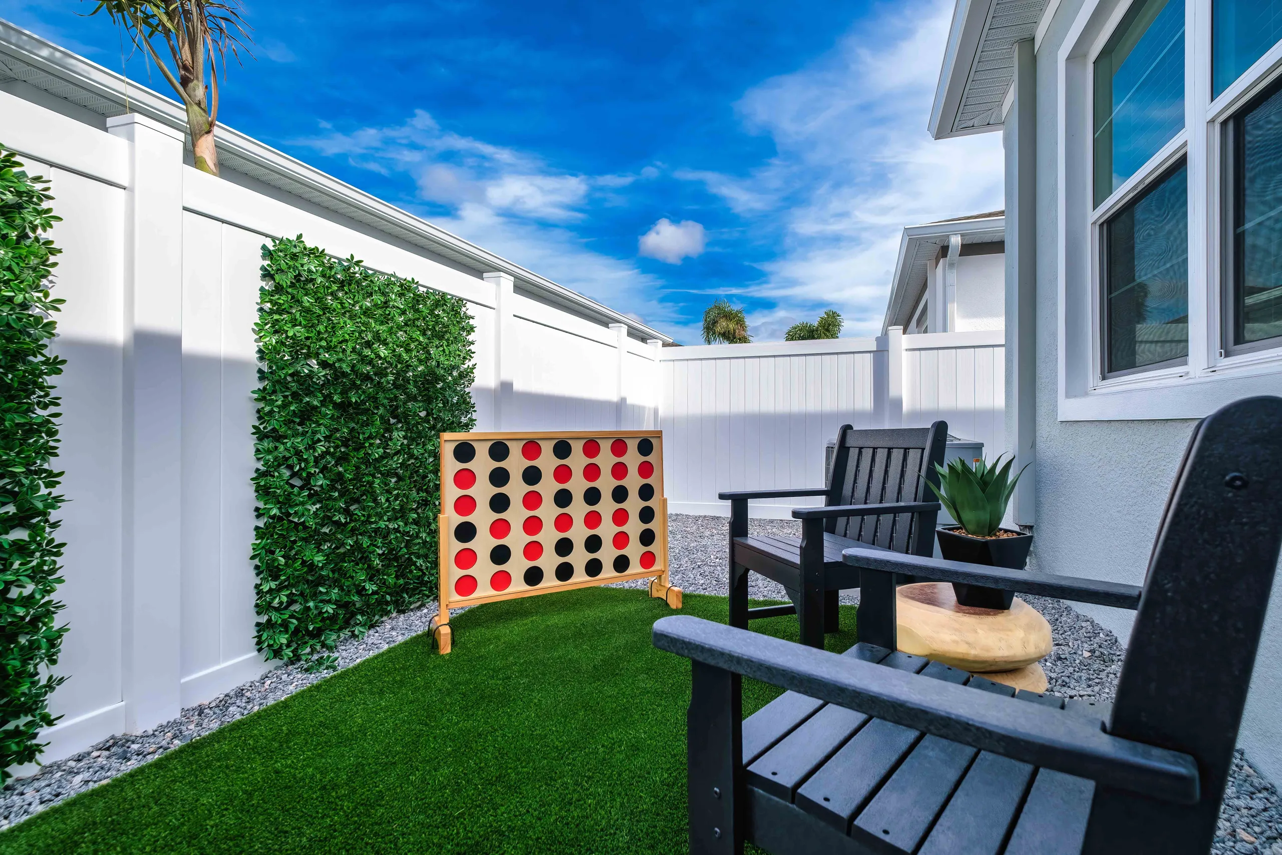 A beautiful white fenced Florida backyard scape with artificial turf, a mature palm tree, seating, a grill, and a patio, on a beautiful breezy sunny day.