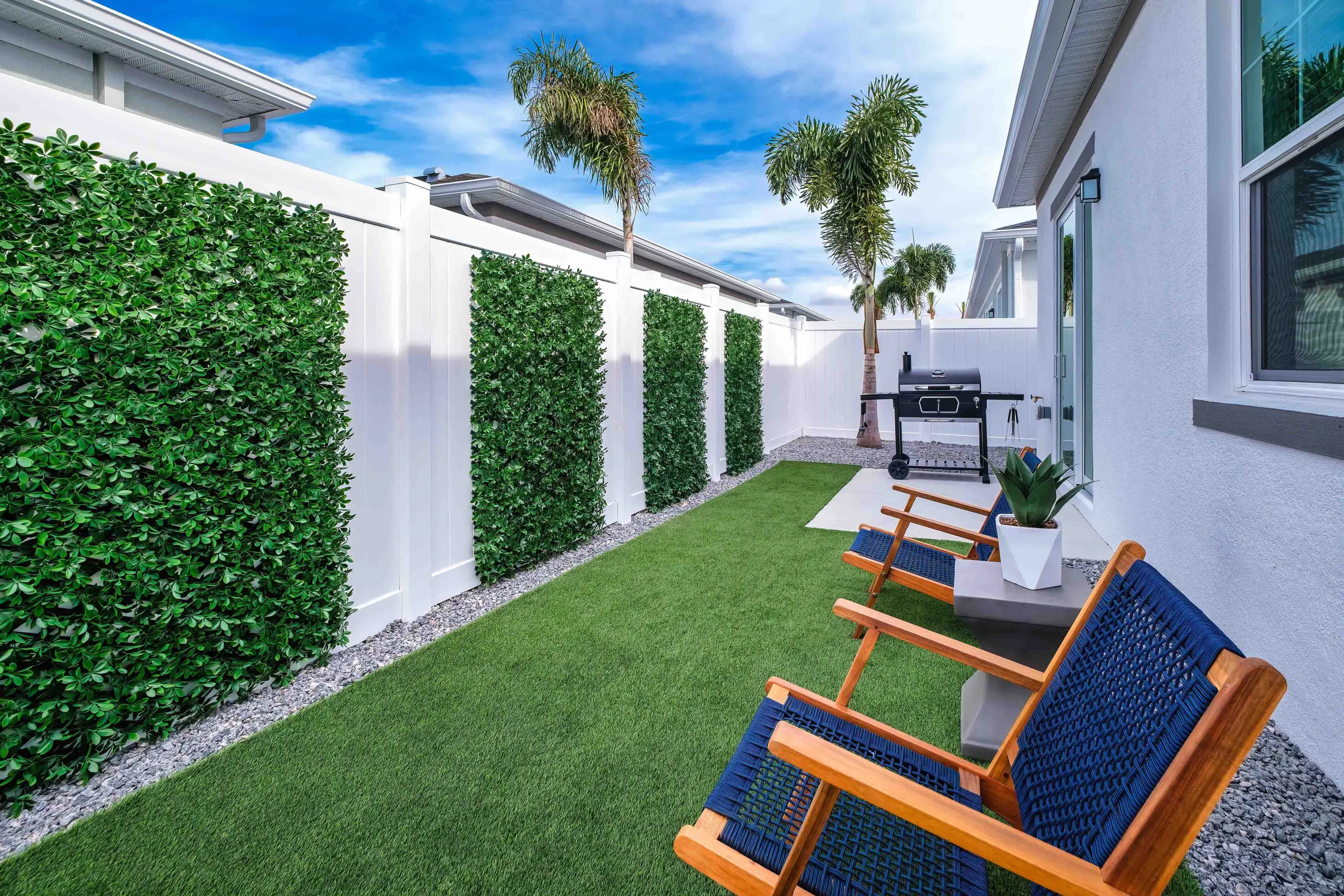 A beautiful Florida backyard scaped with artificial turf, a mature palm tree, seating, a grill, a patio, and a white fence on a beautiful breezy sunny day.