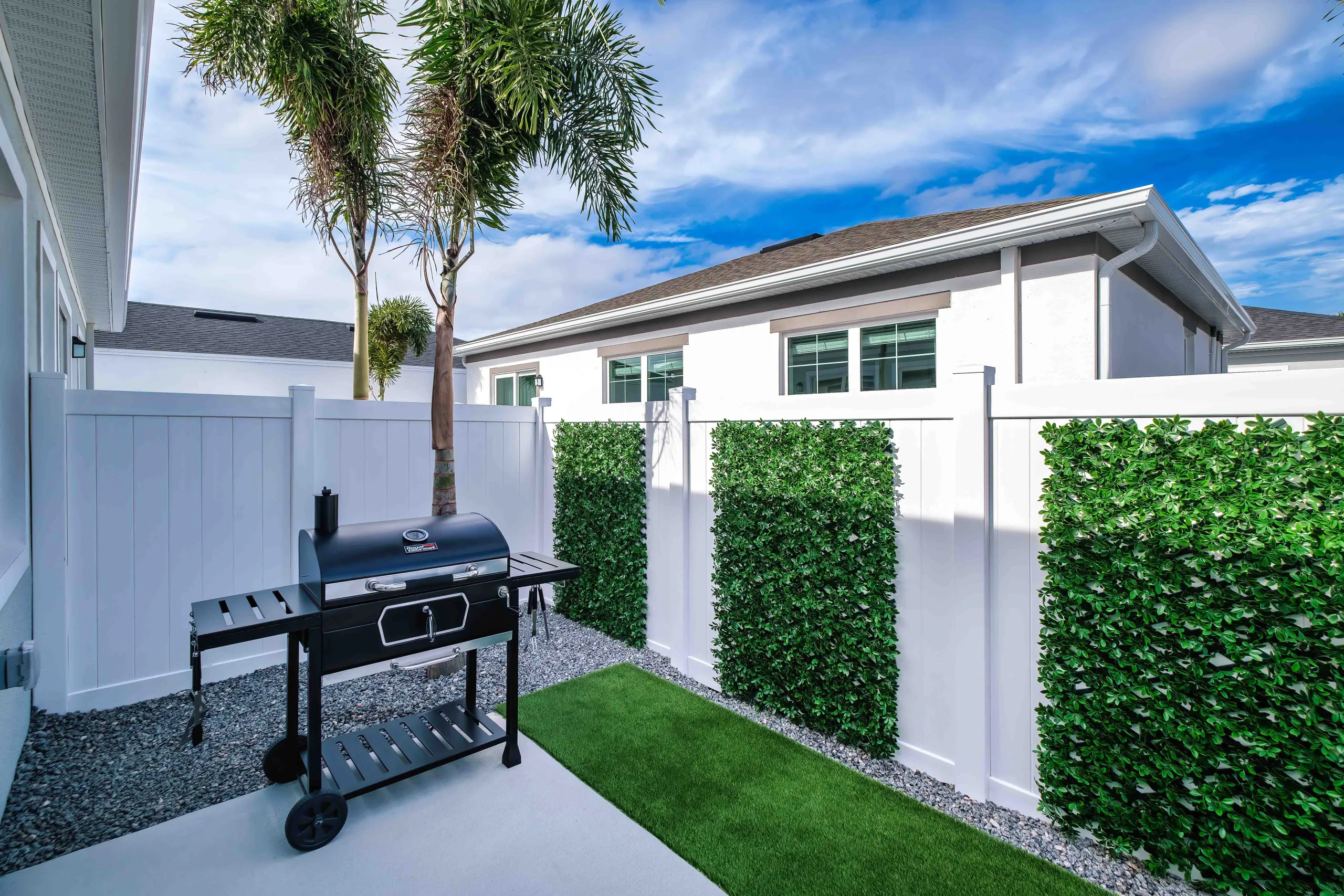 A beautiful white fenced Florida backyard scape with artificial turf, a mature palm tree, seating, a grill, and a patio, on a beautiful breezy sunny day.