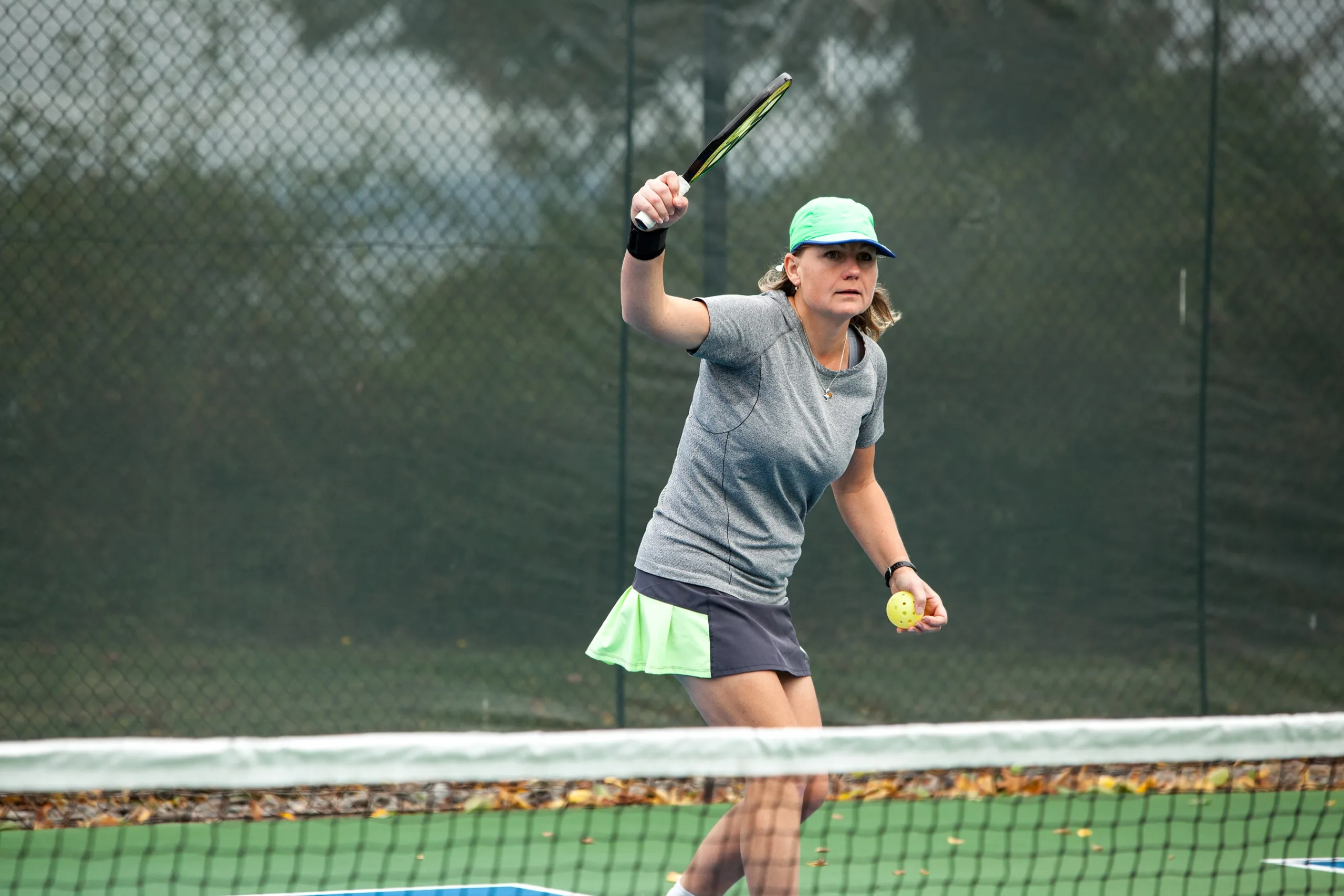 Woman serving a pickleball game wearing a green tennis hat, grey shirt, and black and lime green skirt.