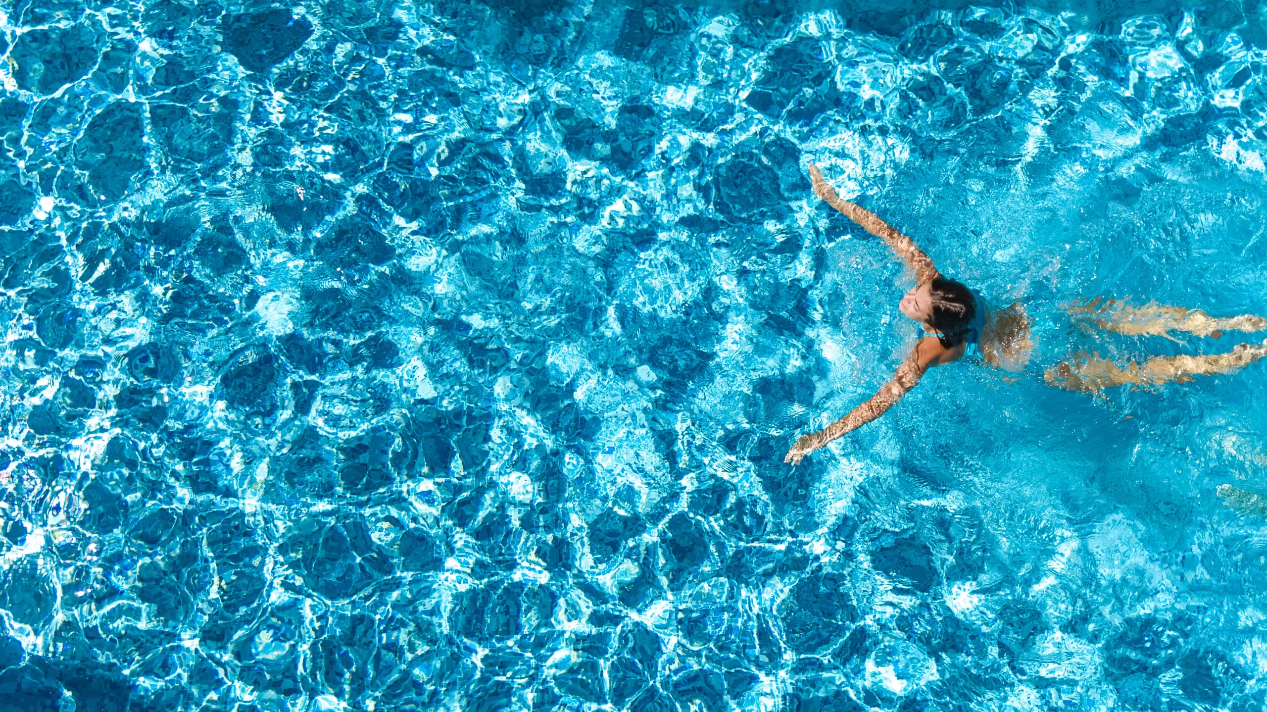 Woman in blue bikini swimming in a pool aerial view photo. She has long brown hair and fair skin.